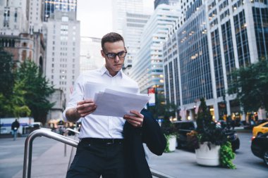 Resmi giyim ve modaya uygun gözlüklü genç işadamı, şehir merkezindeki işlek caddenin arka planındaki parmaklıklara yaslanarak sözleşmenin şartlarını dikkatle araştırıyor.