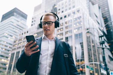 Gözlüklü, neşeli, yakışıklı, yetişkin bir erkekten kablosuz kulaklıkla müzik dinlerken ticari binaların bulanık arka planında akıllı telefonları tarıyor. 