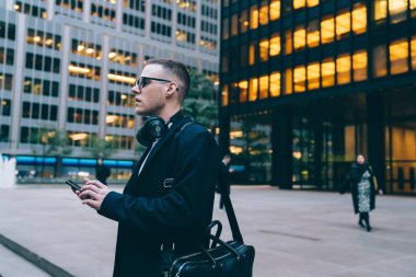 Başarılı ve düşünceli işadamının yan görüntüsü. Parlayan pencereleri olan binaların yanında yürüyen ve şehir caddesine bakmadan akıllı telefonlara bakan.