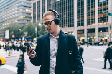 Resmi ceketli ve gözlüklü genç bir girişimci akıllı telefondan mesajları kontrol ediyor ve kulaklıkla müzik dinliyor.
