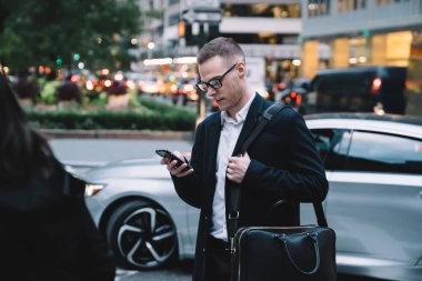 Gözlüklü ve resmi giyinmiş bir adam elinde akıllı telefonuyla karşıdan karşıya geçiyor ve arka planda sosyal medyada gezinirken gülümsüyor.