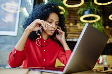 Siyah yetişkin kadın, günlük kıyafetlerle kahve dükkanında masada oturuyor ve bilgisayar başında çalışırken elinde gözlüklerle arama yanıtını bekliyor.