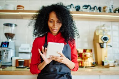 Afro önlüklü, Afro saçlı, genç, çekici bir Afro-Amerikalı kadın. Tablet kullanarak kafede çalışmanın maliyetini hesaplıyor.