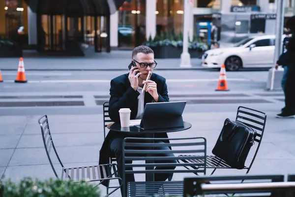 Pensive male entrepreneur in formal attire with glasses talking on smartphone and working on tablet while sitting on downtown street with coffee