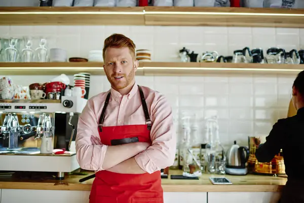Mature redhead male in casual clothes and red apron standing in cafe kitchen with arms folded and looking at camera with smile