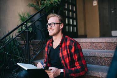 Zeki, gülümseyen, kızıl saçlı, işaretlenmiş kırmızı ve siyah tişörtlü erkek. Not defterine granit merdivenlerde oturan kalemle not alıyor. 