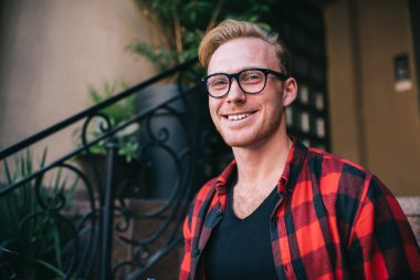 Gülümseyen kızıl saçlı erkek işaretli kırmızı ve siyah tişört ve siyah tişört giyiyor demir çitin yanında duruyor ve kapıya yakın saksılarda bitkiler var. 