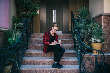 Yaratıcı, kızıl saçlı, gözlüklü, işaretli kırmızı ve siyah tişörtlü bir erkek. Yazın şehirde granit merdivenlerde oturan kalemle not alıyor. 