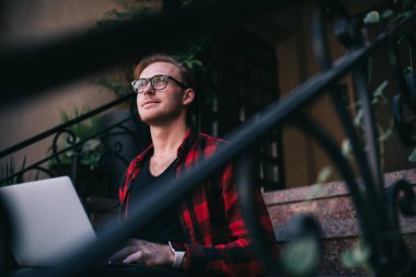 Ekose gömlekli ve gözlüklü genç adam dizüstü bilgisayarını kullanıyor. Projeyi düşünüyor. Merdivenlerden yukarı bakıyor.