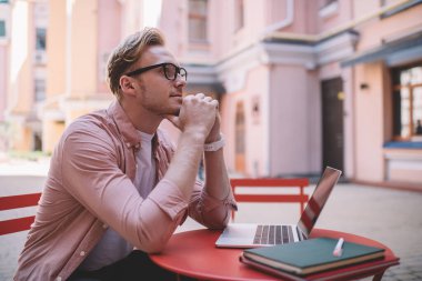 Gözlüklü genç düşünceli bir adamın, kafenin dışında dizüstü bilgisayarla otururken eline yaslanıp ilham arayışının yan görüntüsü.