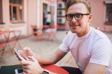 Keyifli gülümseyen adam günlük giysiler ve gözlük takıyor. Yazlık kafede video izlemek için tablet kullanıyor. Kameraya bakıyor.