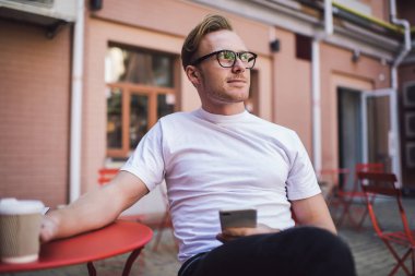 Düşünceli genç adam günlük giysiler ve bardaklar içinde bir fincan kahve alıp sokak kafesinde sosyal medyada gezinip gözlerini kaçırıyor.