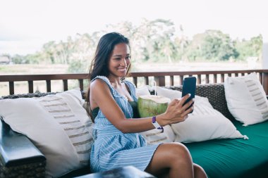 Neşeli yetişkin Asyalı kadın akıllı telefonuyla selfie çekerken balkonda taze hindistan cevizi içeceği ve bulanık yeşil tropikal ormanlara karşı rahat bir kanepede oturuyor.