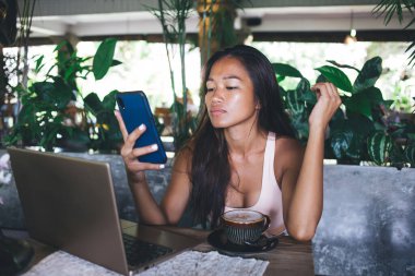 Kendine güveni tam bir etnik kadın, kafeteryada dizüstü bilgisayarı ve kahve fincanı ile oturmuş akıllı telefondan internette sörf yapıyor, elini kaldırıyor ve göğüs dekoltesini gösteriyor.