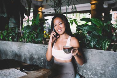 Neşeli genç Asyalı kadın, beyaz bir bluz giyip restoranda cappuccino içerken cep telefonuyla kameraya bakarak arama yapıyor.