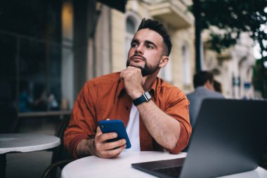 Düşüncelere dalmış düşünceli genç bir erkek. Kafe terasında dizüstü bilgisayarla oturmuş cep telefonuna bakarken başını çeviriyor.