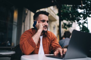Gündelik giysiler içinde gülümseyen genç bir erkek müşteri kafede otururken bilgisayarını karıştırırken keyifli bir şekilde telefonla konuşuyor.
