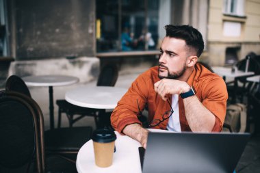 Gündelik giyinmiş, akıllı bir öğrenci dizüstü bilgisayarda çalışıyor ve kafeteryada kahve fincanı ile otururken dalgın dalgın bakıyor.