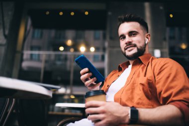 Aşağıdaki neşeli ve iyimser adamın sokak kafesinde oturup sosyal medyada mesajlaşırken gülümsemesi ve kameraya bakması.