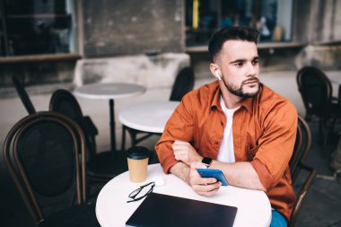 Elinde akıllı telefon olan sakallı bir adam kafede oturup kulaklıkla müzik dinliyor ve bulanık arka plana bakıyor. 