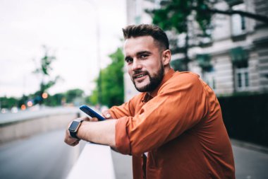 Yakışıklı, sakallı, sıradan turuncu tişörtlü, akıllı telefon kullanan, çite dayanmış kameraya bakan hippi bir adamın portresi.