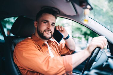 Pahalı bir arabada oturan, direksiyonu çevirirken akıllı telefonla konuşan ve arka plandaki bulanık kameraya bakan günlük kıyafetli erkek sürücünün yan görüntüsü.