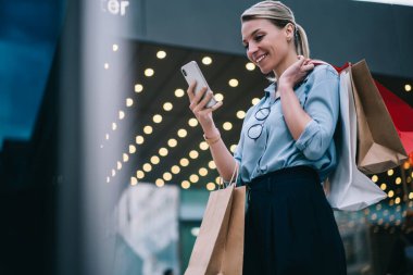 Gülümseyen kadın gözlüklü müşteri dışarıda alışveriş torbalarıyla dikiliyor ve akıllı telefon cihazında kablosuz internet kullanıyor, arama yerleri için cep telefonu uygulaması kullanıyor.