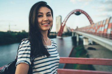 Gülümseyen Asyalı hippi kız gezginin yarım boy portresi. Yaz tatilinin tadını çıkarıyor. Mutlu genç hippi kız nehir köprüsünde kameraya bakıyor.