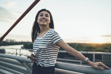 Gezinin tadını çıkaran gülümseyen Asyalı gezgin elinde cep telefonu olan şehir köprüsünde duruyor, mutlu kadın blogcu akıllı fonla dolaşırken 4G internet bağlantısı üzerinden medya içeriğini paylaşıyor