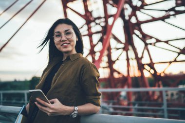 Neşeli giyinmiş kadın blog yazarının yarım boy portresi. Şehir köprüsünün arka planında elinde akıllı telefon duruyor. Asyalı hippi kız cep telefonuyla cep telefonundan uygulamayla kameraya bakıp sohbet ediyor.