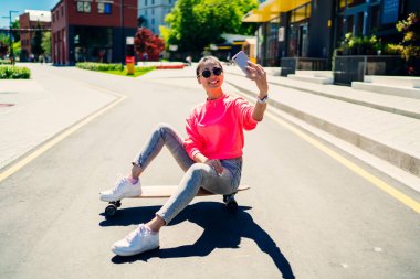 Asyalı kadın etkileyicinin takipçilerle uzun kaykay sporu hakkında iletişim kurmak için video bloğu çekmesi ve akıllı telefon kamerasına gülümsemesi, milenyum blog yazarının selfie çekmesi.