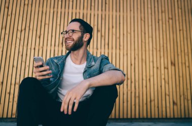 Şık sokak kıyafetleri giymiş başarılı bir beyaz adam cep telefonuyla iletişim kurarken şehirdeki 4G kablosuz bağlantısını kullanıyor. Neşeli hipster adam blog sitesi.