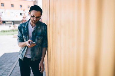 20 'li yaşlardaki modaya uygun giyinmiş blogcu, cep telefonunda internet sitesine bağlanmak için klasik gözlüklerle güncellemeye odaklanmış akıllı telefon bildirimini kontrol ediyor.