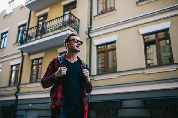 Low angle of handsome pensive man in sunglasses and black and red casual clothes with backpack looking away and dreaming while standing alone on street in city