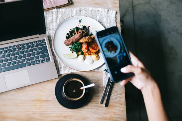 Faceless person in restaurant taking photo of plate with food on table with gray laptop, coffee cup, cutlery and tablecloth