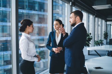 Gündüz vakti şehir konferans odasındaki pencerenin önünde birlikte dururken resmi kıyafetli başarılı iş arkadaşları konuşuyor ve el kol hareketi yapıyor.