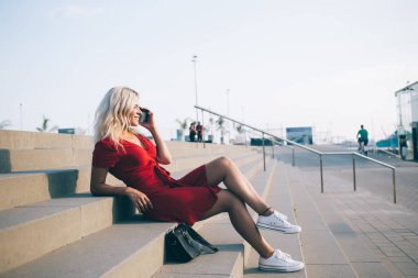 Kırmızı elbiseli, çekici, neşeli bir kadının cep telefonuyla konuşurken kenar mahallelerdeki merdivenlerde otururken kendinden emin bir şekilde bakışları başka yöne bakıyor.