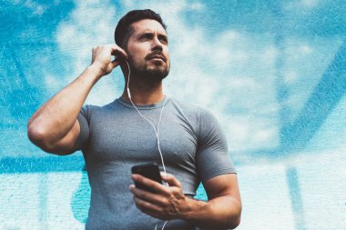 Spor kıyafetli, kulaklık takan, cep telefonuyla müzik dinleyen, sokakta koşmaya hazırlanan ve gözlerini kaçıran kaslı bir adamdan.