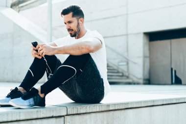 Spor kıyafetleri içinde, kulaklıklı yakışıklı, gezici sosyal medyada müzik mesajları dinleyen merdivenlerde oturan yakışıklı adam.