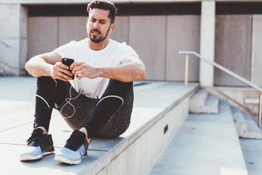 Aktif görevde olan sakallı genç sporcu, şehir caddesindeki sallanan merdivenlerde bacakları bükülmüş halde otururken cep telefonu ve kulaklık takıyor. 