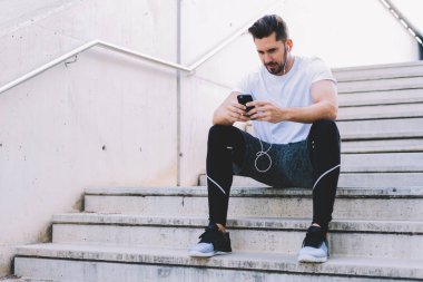 Spor kıyafetleri içinde sakallı genç bir erkek atlet binanın merdivenlerinde oturuyor ve kulaklıkla iletişim kuruyor. 
