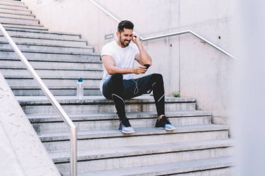 Aktif giyinen ve spor ayakkabı giyen neşeli erkek sporcu şehir caddesindeki binanın merdivenlerinde dinlenirken kulaklıkla cep telefonuyla video görüşmesi yapıyor. 