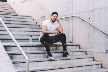 Düşük açılı sakallı sporcu aktif giyimli ve spor ayakkabılı tren için hazırlanırken kulaklık takıyor ve şehir caddesinde cep telefonu kullanıyor. 