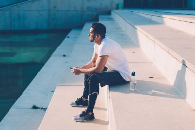 Spor kıyafetli, kulaklıklı, yürüyüş sonrası dinlenen telefonları olan, merdivenlerde oturan ve gözlerini kaçıran düşünceli sakin bir erkeğin yan görüntüsü.