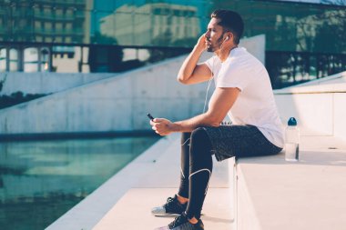 Kulaklıklı, kulaklıklı, kulaklıkları kontrol eden, müzik dinleyen ve şişeyle aydınlık bir günde merdivenlerde dinlenen aktif sakallı sporcuların yan görüntüsü. 