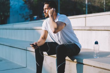 Dalgın içerikli sporcunun kulaklıklara dokunuşunun ve cep telefonuyla sörf müziğinin parlak bir günde merdivenlerde oturuşunun yan görüntüsü 