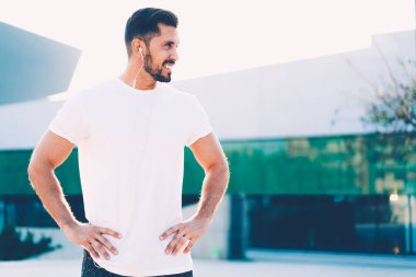 Güçlü, güçlü, gülümseyen, kulaklıklı, müzik dinleyen ve elleri bir yana, cam bina boyunca bakan koşucunun yan görüntüsü. 