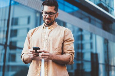 Gündelik giysiler ve gözlüklerle gülümseyen başarılı adamdan modern şehrin sokaklarında sosyal medyada gezen moible mesajlar kullanarak.