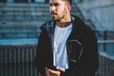 Ciddi yetişkin bir erkek, elinde cep telefonu olan beyaz gömlekli ve siyah ceketli. Kulaklıklı bir radyo dinliyor, şehirde dikiliyor ve gözlerini kaçırıyor.