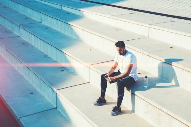 Spor kıyafetleriyle merdivenlerde oturmuş, akıllı telefon ve kulaklıkla müzik dinlerken şehirdeki eğitimden sonra dinlenen aktif bir erkek. 
